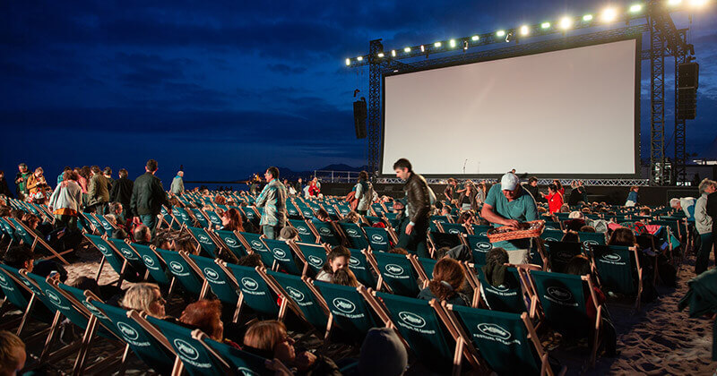 Cannes Film Festival outdoor screening. People seated in chairs on a beach watch a large screen at night. Film submissions.